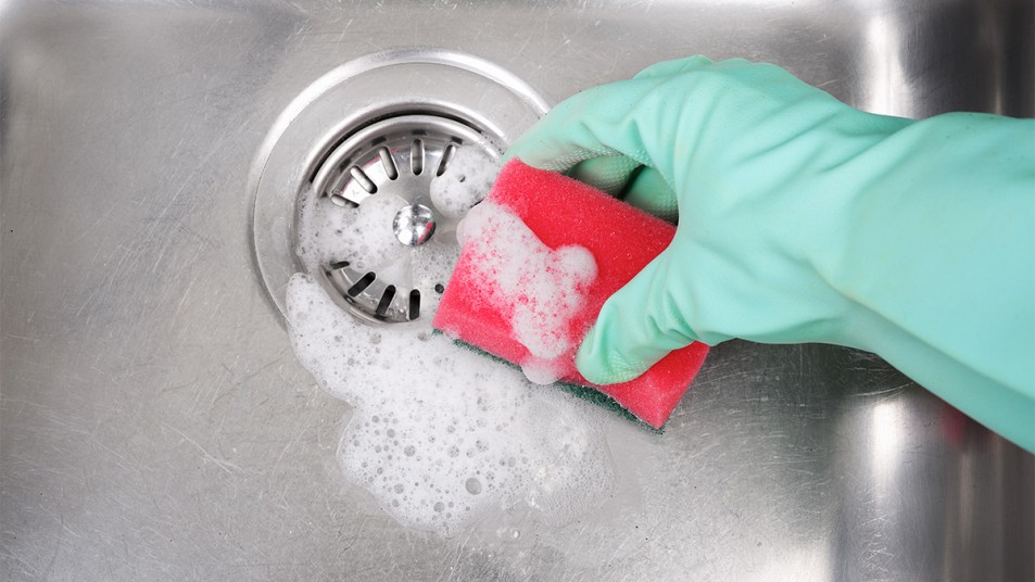 hand wearing a glove cleaning a stainless steel sink with a sponge