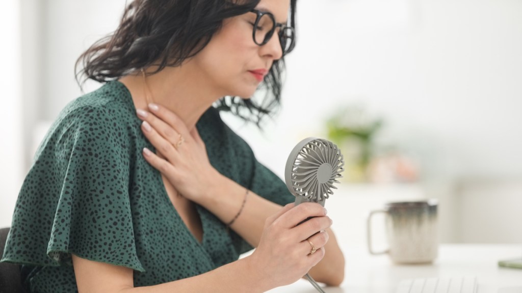 A woman with dark hair holding her hand to her chest while she blows a fan on herself to cool a hot flash