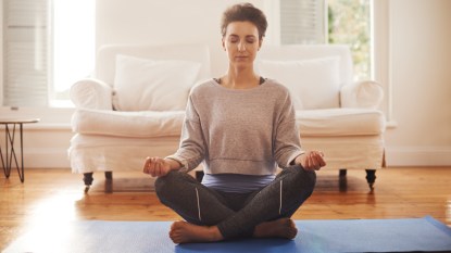 A woman sitting on a mat in her living room with her hands on knees and eyes closed practicing yoga for menopause