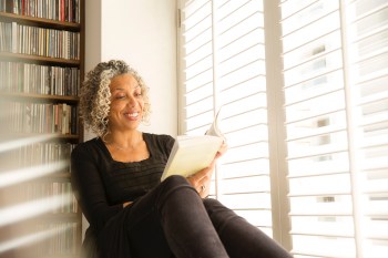 older black woman reading a book while leaning against a bookshelf