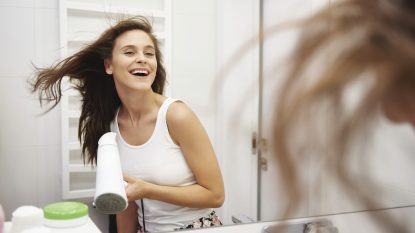 Mirror image of laughing woman blow-drying her hair in the bathroom