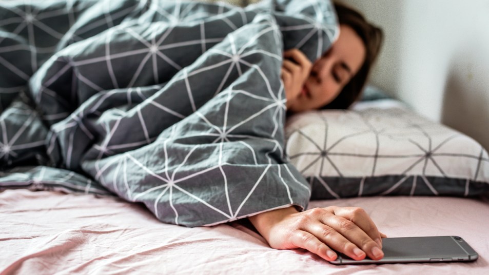 Woman under bed covers reaching for her phone