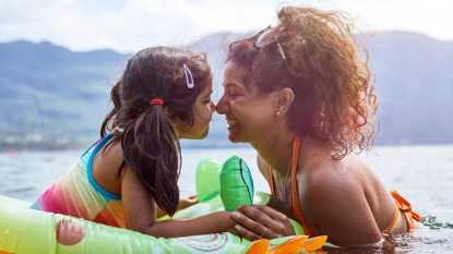 Mom with young daughter in a floatie swimming in a lake surrounded by nature