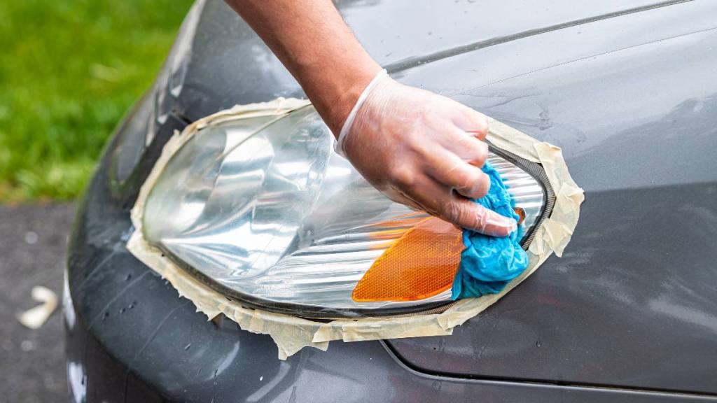 Uses for toothpaste: worker cleaning car headlights taped to protect paint rubbing with blue shop towel closeup on driveway of house outside 