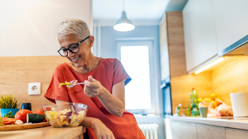 mature woman with short gray hair and glasses, smiling, eating food from a bowl in the kitchen