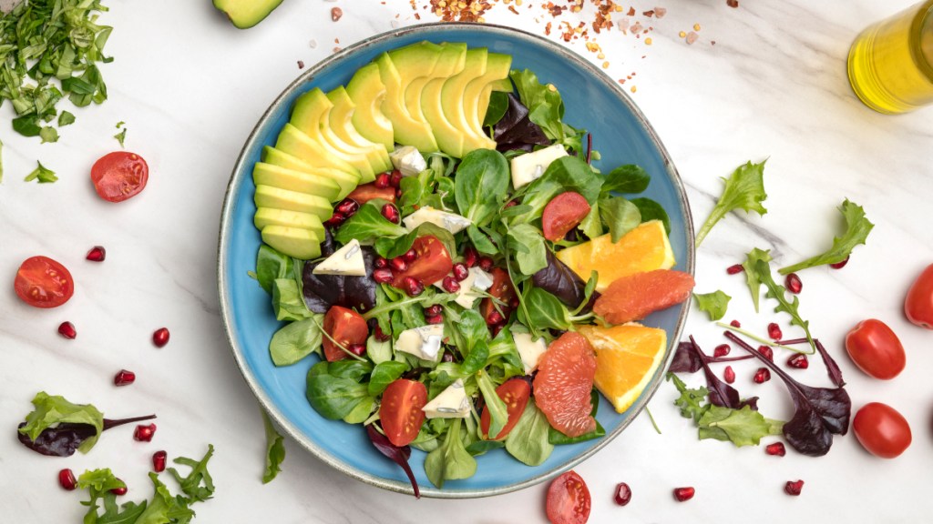 A watercress salad, which has many health benefits, topped with avocado, oranges, pomegranate seeds, tomatoes and cheese on a blue plate