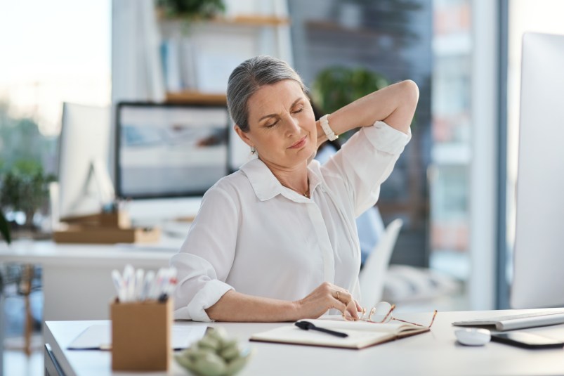 mature woman in white t-shirt sitting at desk, rubbing shoulders in pain