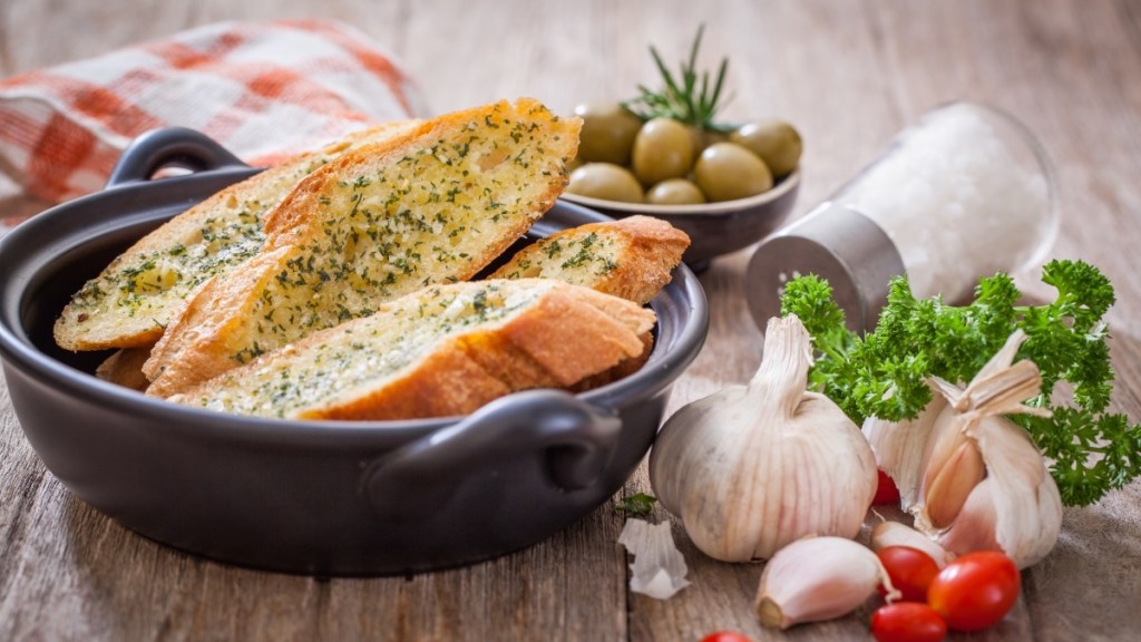 A dish of garlic bread, which can help get rid of a UTI in 24 hours, beside fresh garlic, olives and cherry tomatoes