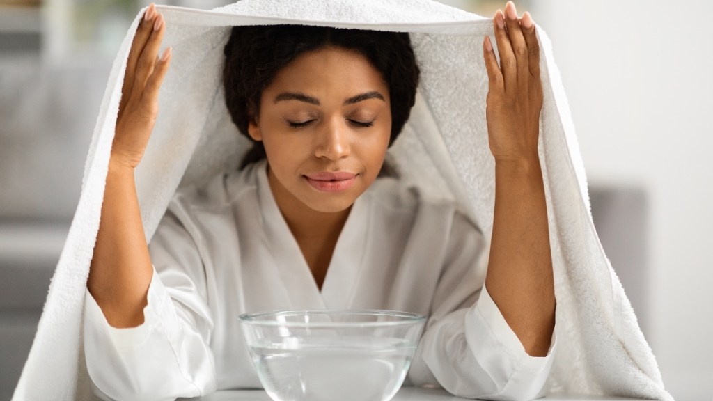 A woman in a white robe using a white towel over head to trap the steam from the bowl of eucalyptus essential oil and water on the table