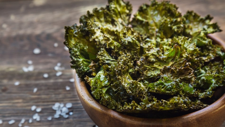 Air fryer kale chips in a wooden bowl on a wooden table covered in salt sprinkles