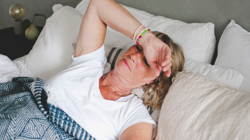 Mature white woman lies in bed with her arm over her face.