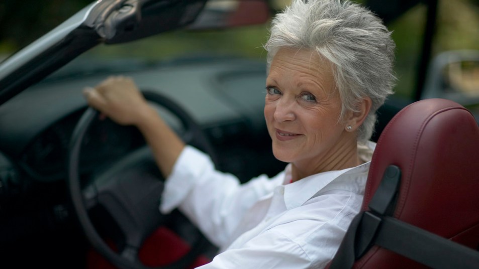 Mature woman with a short white haircut sits at her steering wheel smiling and wearing a white shirt.