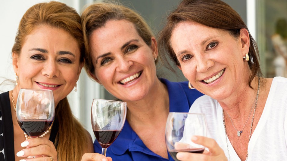 Three white, middle-aged women hold up their wine glasses and smile for the camera.