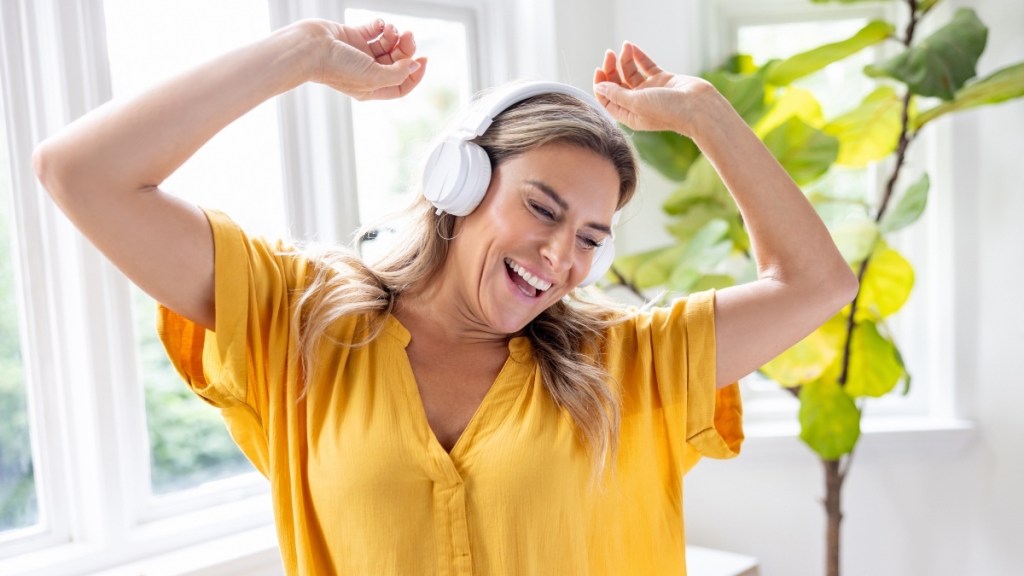 A woman in a yellow top wearing white headphones with her hands raised, dancing, to increase serotonin