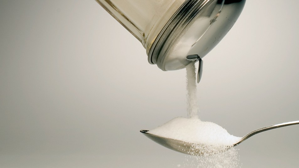 pouring sugar from a glass jar into a spoon against a gray backdrop