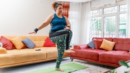 mature woman with curly hair wearing blue-green workout clothes, doing moderate intensity exercise in front of yellow and red couches
