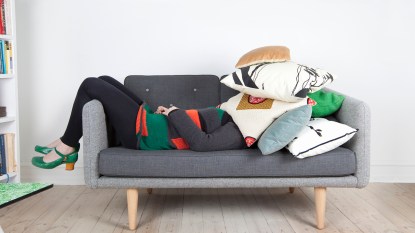 Woman lying on sofa with pile of pillows over her head.
