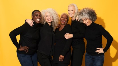 A group of mature women join in a happy, laughing huddle against a golden yellow background.