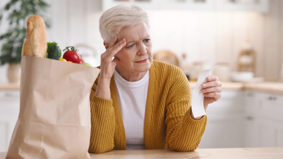 senior woman upset looking at receipt, groceries on table, inflation spike concept