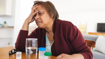 mature woman suffering from a migraine, head in her hand, with pills on the table next to a glass of water