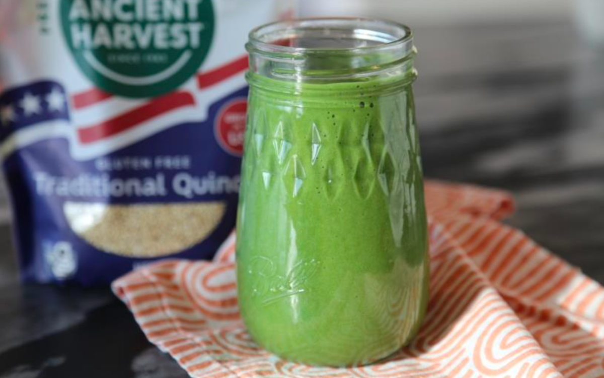 green power smoothie in a clear glass with a bag of Ancient Harvest quinoa behind it
