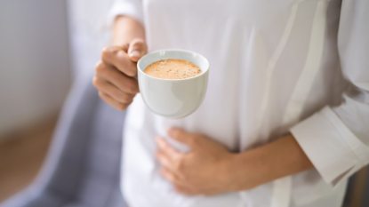 mature woman holding cup of coffee in one hand, other hand holding upset stomach