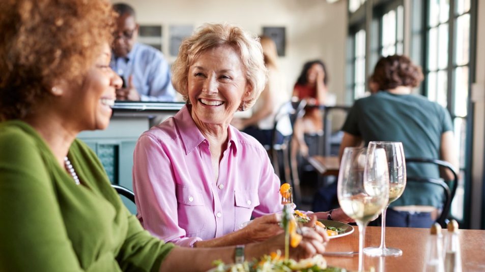 mature woman in green shirt and mature woman in pink shirt smile as they eat restaurant food