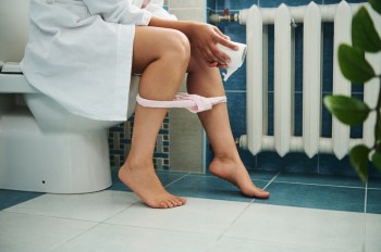 Cropped view of woman in bathroom sitting on toilet with toilet paper in hand.