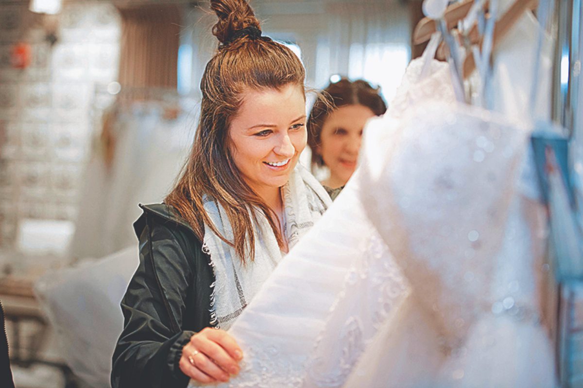 woman with high bun and black jacket smiling as she looks through a rack of wedding dresses