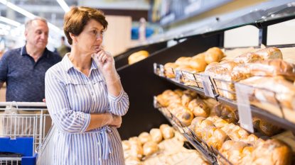 mature woman choosing a loaf of bread in the grocery store, husband behind her