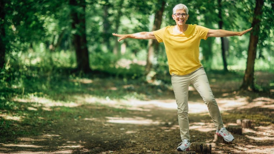 mature woman outside, smiling, balancing on one foot on a log