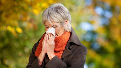 mature woman suffering from fall allergies, blowing her nose outside wearing orange scarf
