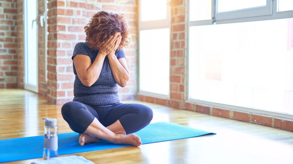 mature woman exercising on yoga mat sitting, covering face, bladder leaks concept