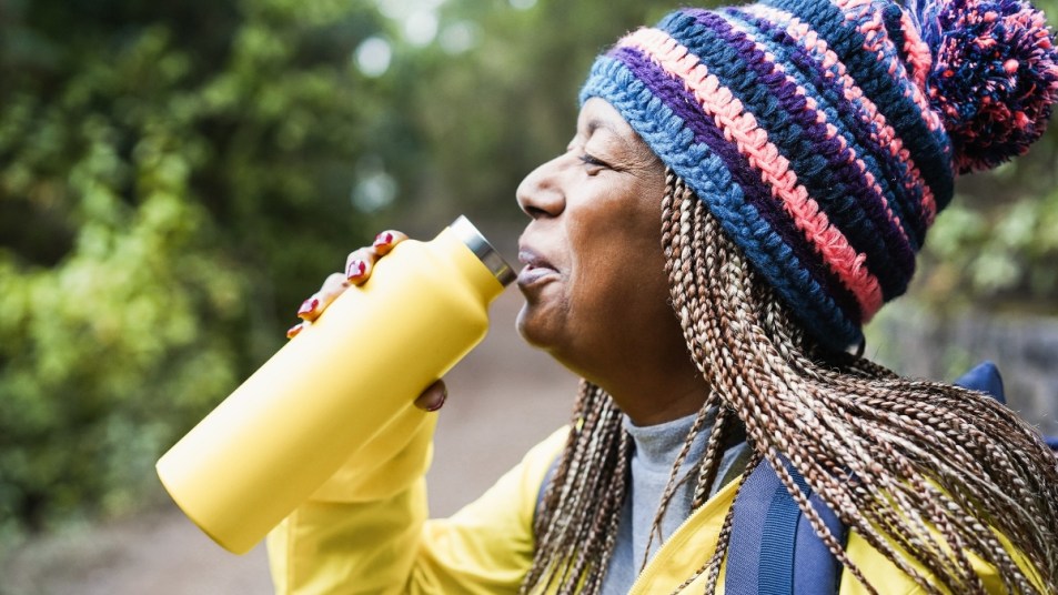 Senior african woman drinking water during trekking day in mountain forest - Focus on hat