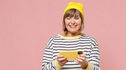 Elderly fun woman 50s in striped shirt yellow hat using play racing app on mobile cell phone hold gadget smartphone for pc video games isolated on plain pastel light pink background studio portrait.
