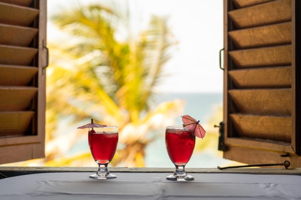 Two sorrel rum punches resting on a table with a view to coconut trees and the ocean outside