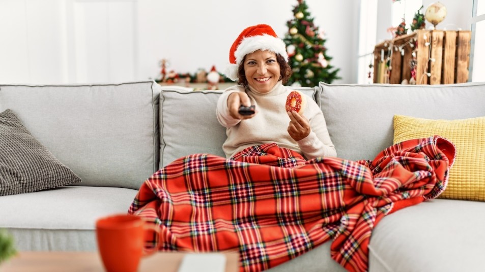 Woman wearing Santa hat and holding remote while sitting on couch under plaid blanket