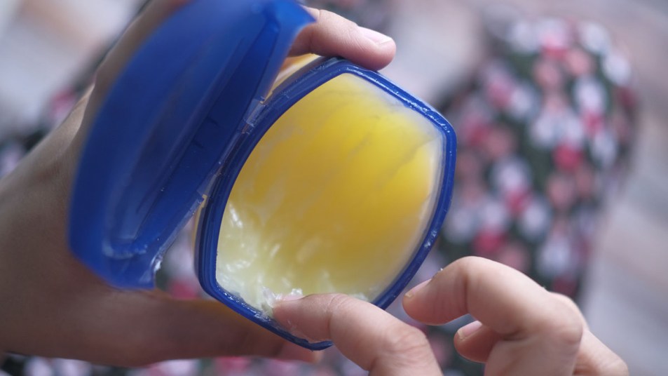 A pair of hands hold an open jar of petroleum jelly and a finger is shown dipping into it.