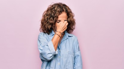 Middle age beautiful woman wearing casual denim shirt standing over pink background tired rubbing nose and eyes feeling fatigue and headache. Stress and frustration concept.
