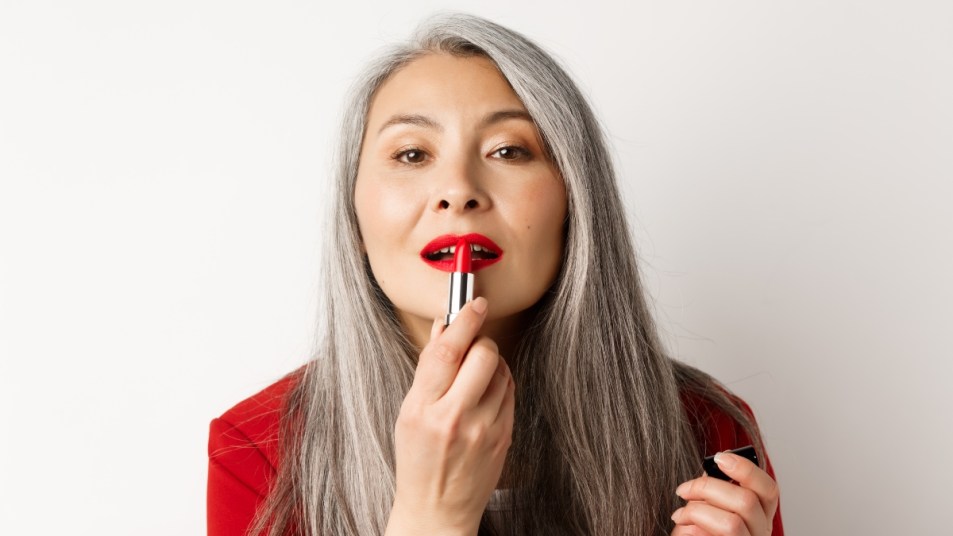 Beauty and fashion concept. Close up of senior asian woman looking in mirror and apply red lipstick, standing over white background