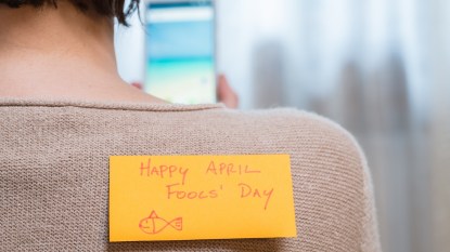 Woman looking at her phone with "Happy April Fools' Day" post-it on her back