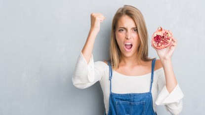 Beautiful young woman over grunge grey wall holding pomegranate annoyed and frustrated shouting with anger, crazy and yelling with raised hand, anger concept