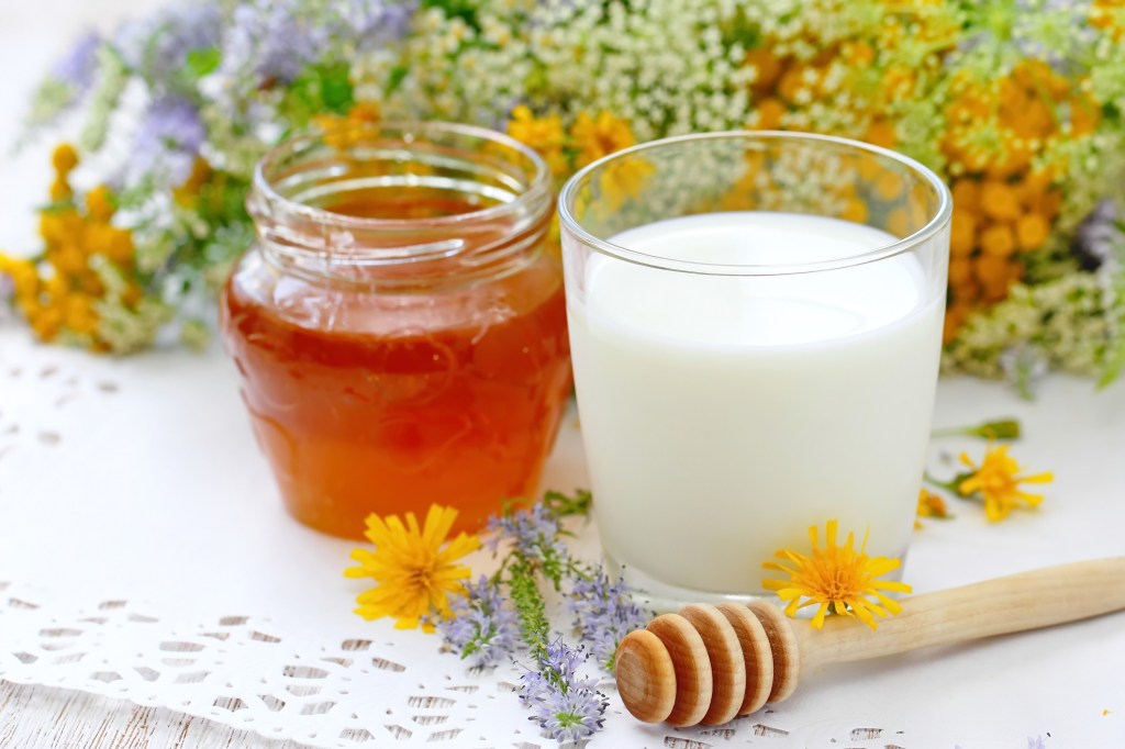 Photo of a glass of milk and fresh honey in jar with flowers background for use in DIY hand soak to relieve dry, cracked skin