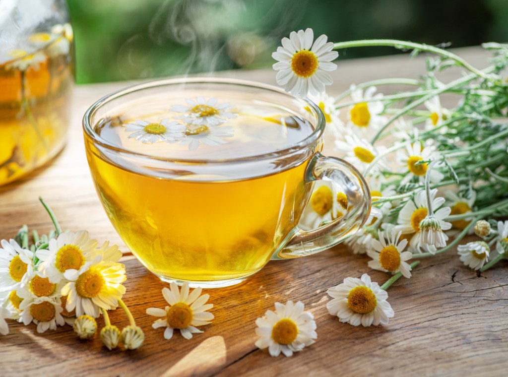 A photo of a mug of warm chamomile tea on a table surrounded by chamomile flowers to be used in a soothing hand soak for dry skin