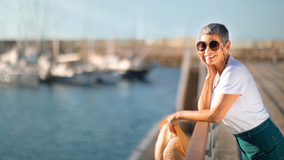Dream Vacation. Happy Senior Lady Posing With Sunglasses Holding Summer Straw Hat, Standing At Sea Pier With Yachts. Relaxed Tourist Woman Enjoying Holidays And View At Marina Dock.
