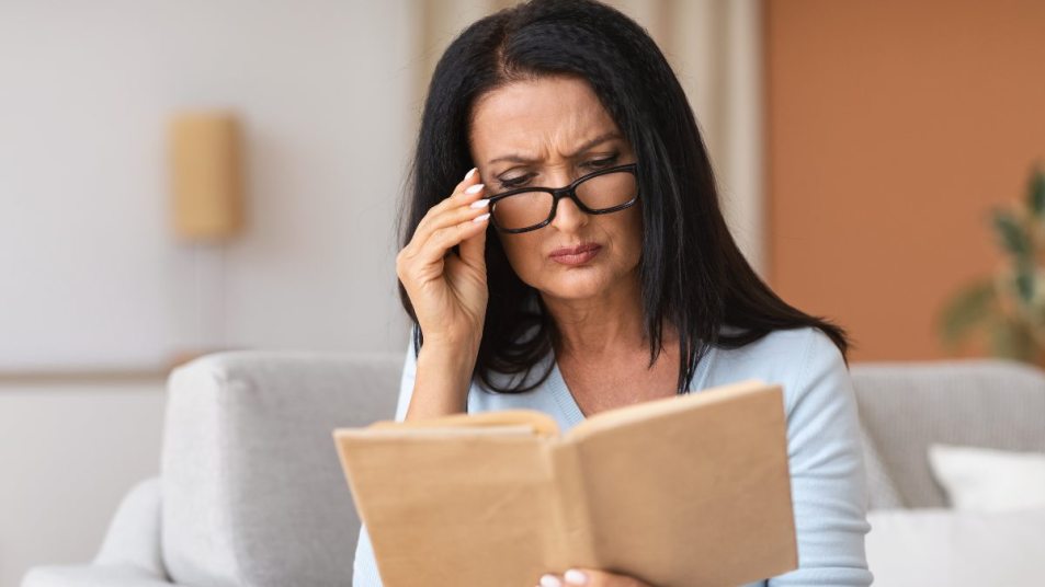 mature woman with black hair struggling to read, hand to her glasses, book in hand