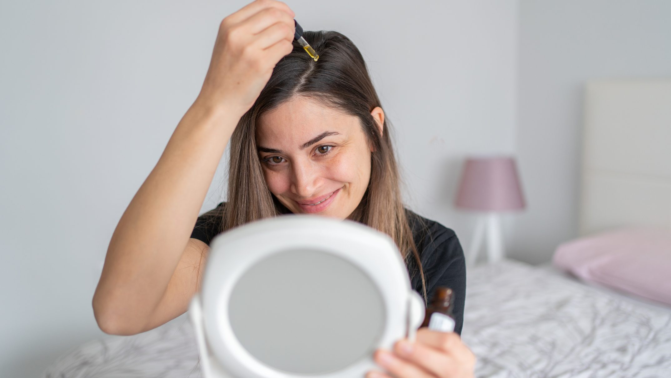 Woman applying oil to scalp in mirror.
