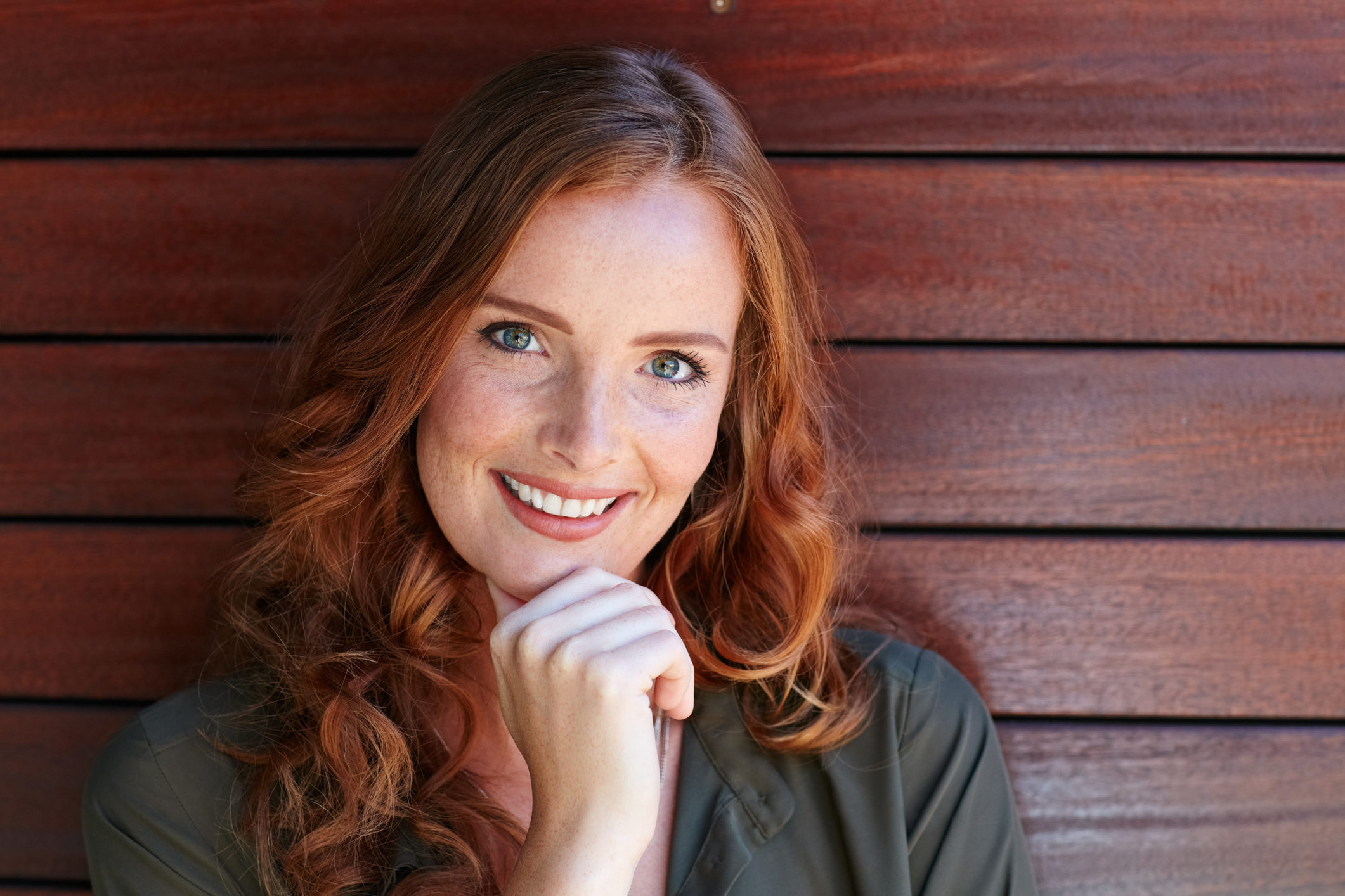 Woman with red hair posing in front of a wood wall while smiling and touching her chin