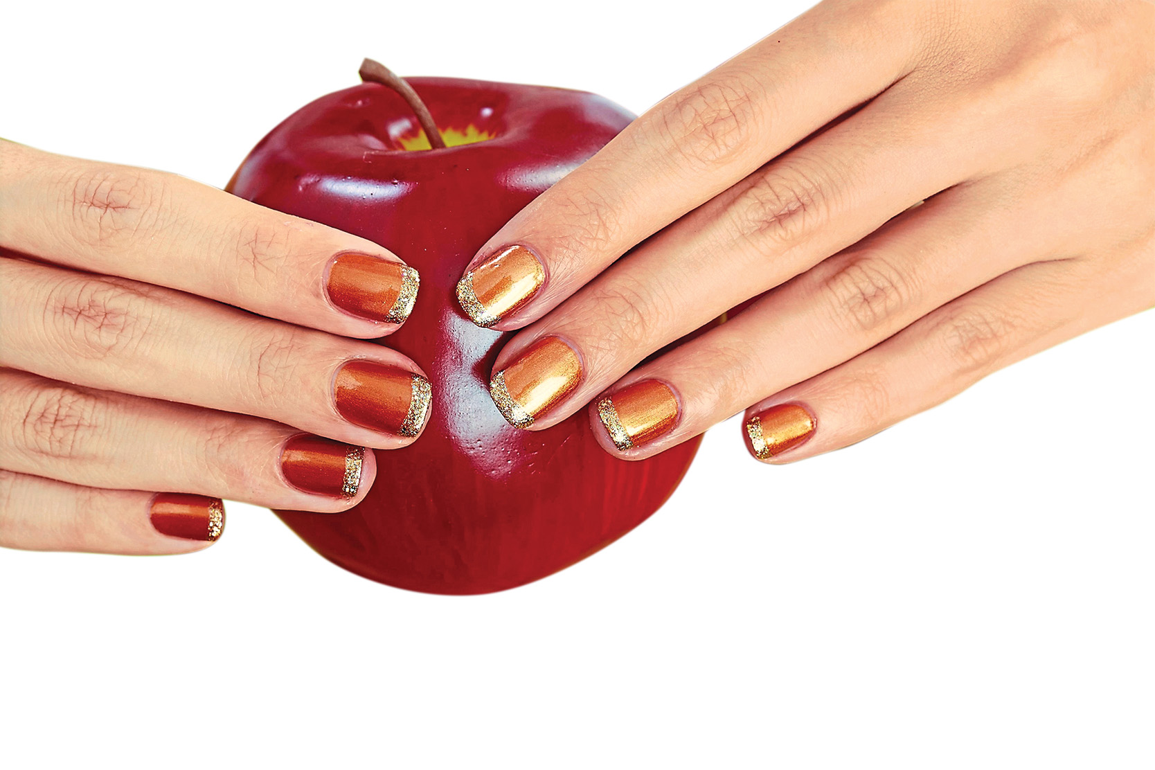A woman holding an apple with nails painted with a French manicure in orange and gold polish shades