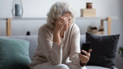 mature woman stressed, sitting on couch, head in hand, looking at her phone, technology issues